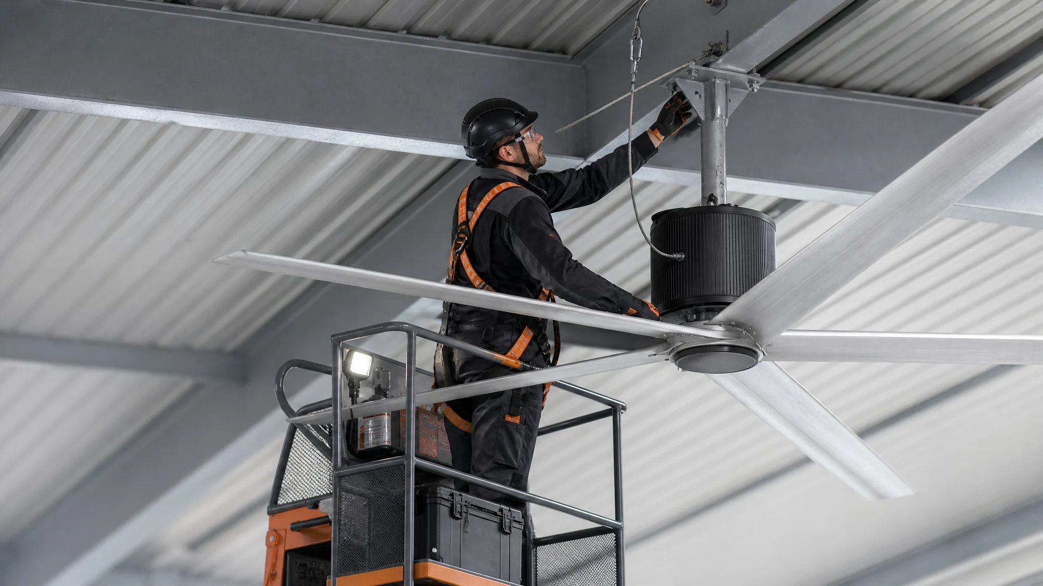 Technician inspecting an HVLS fan mounting bracket, safety retention cable, motor housing, hub and six airfoil roots from a secured lift platform